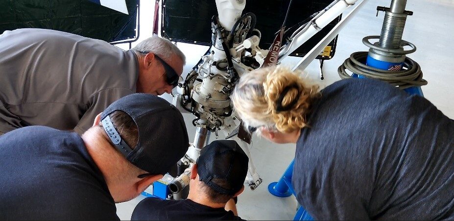 Four people gathered closely around the main landing gear of a business jet in a hangar, being shown the component at close range.